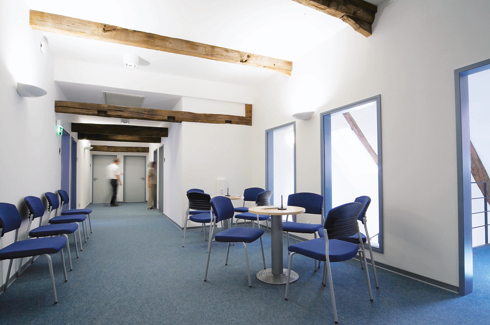 Moderner Wartebereich mit blauen Stühlen, Holzbalken an der Decke und zwei Personen im Hintergrund.