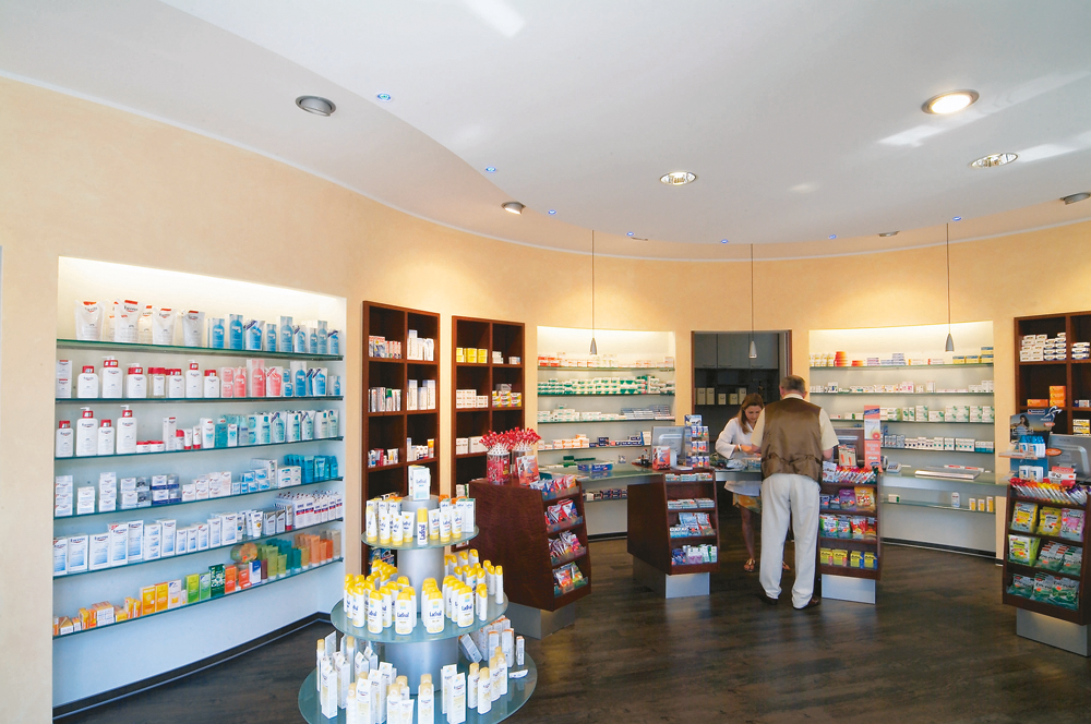 Innenansicht einer Apotheke mit Regalen, Präsentationstisch und zwei Personen am Verkaufstresen.
