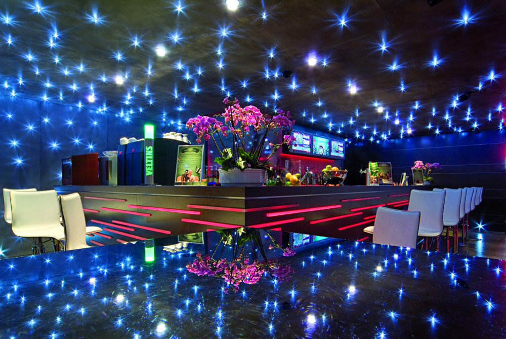 Moderne Bar mit polierter Oberfläche, weißen Hockern, rosa Orchideen und blauer sowie roter Beleuchtung.