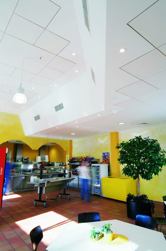 Moderne Kantine mit optimierter Akustikdecke für Schallschutz, großen weißen Deckenelementen mit integrierter Belüftung, warm gestalteten Wänden in Gelb- und Orangetönen, einem Buffetbereich und gemütlicher Sitzgelegenheit mit Pflanzen.