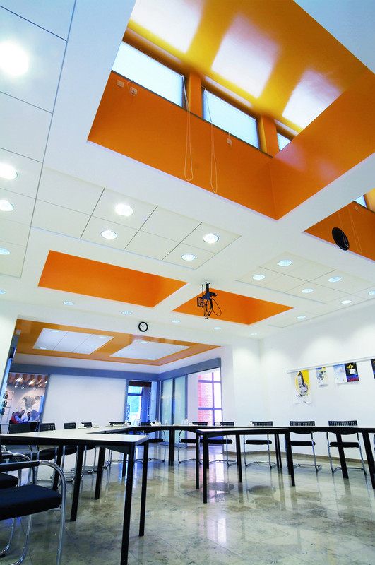 Modern gestalteter Konferenzraum mit Schallschutzdecken in weiß-orange, integrierter Beleuchtung und Projektionstechnik, ausgestattet mit schwarzen Stühlen und Tischen für Meetings oder Schulungen.