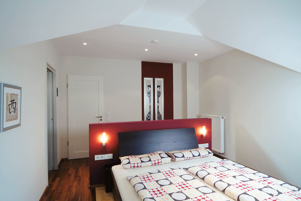 Modernes Schlafzimmer mit großem Bett, bunter Bettdecke, Wandleuchten und abstraktem Bild an roter Wand.
