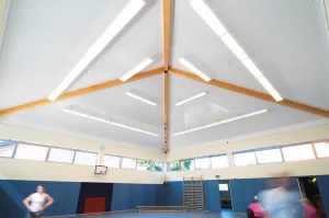 Moderne Turnhalle mit Holzbalkendecke, blauen Matten, Sprossenwand und unscharfen Personen in Bewegung.