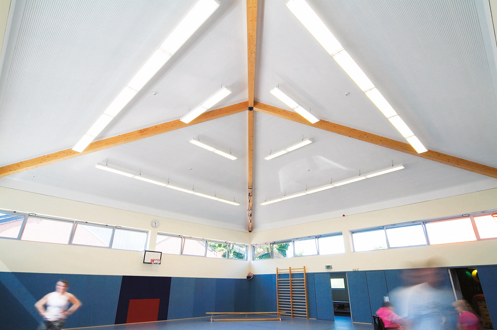 Moderne Turnhalle mit Holzbalkendecke, blauen Matten, Sprossenwand und unscharfen Personen in Bewegung.