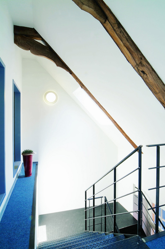 Eine modernisierte Treppe mit schwarzem Geländer und blauen Teppichböden. Sichtbare alte Holzbalken verleihen dem Raum einen rustikalen Charme, während die weißen Wände und großen Fenster für eine helle Atmosphäre sorgen.