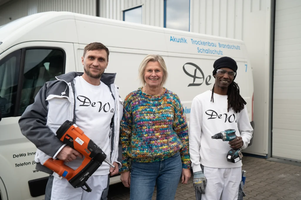 Zwei Handwerker von DeWa Innenausbau mit einer Frau vor einem Firmenfahrzeug, ausgestattet mit Werkzeug und Arbeitskleidung, bereit für den nächsten Einsatz.