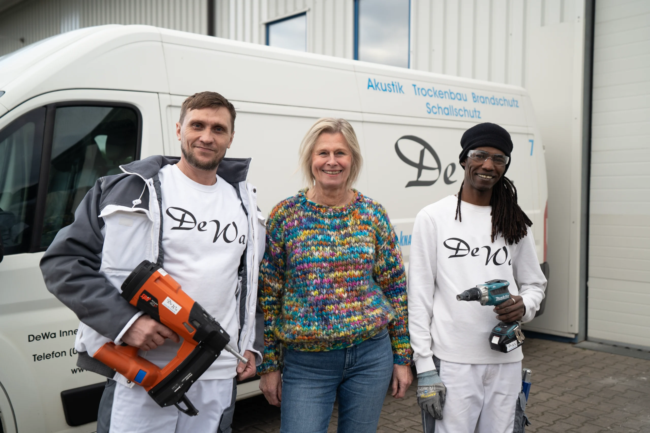 Zwei Handwerker von DeWa Innenausbau mit einer Frau vor einem Firmenfahrzeug, ausgestattet mit Werkzeug und Arbeitskleidung, bereit für den nächsten Einsatz.