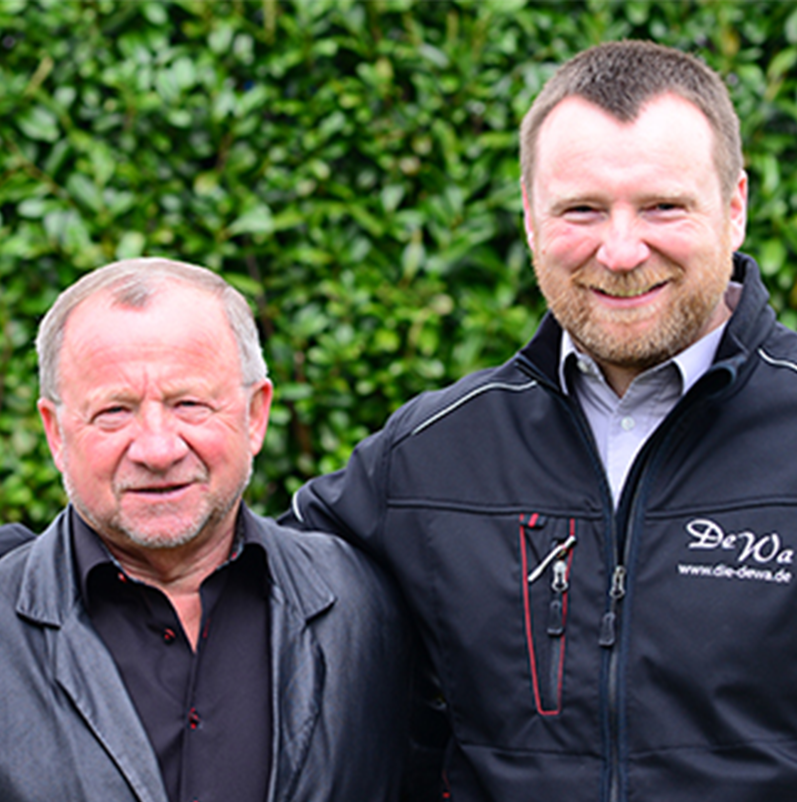 Alfred und Ralf Müller lächelnd vor einer Hecke.
