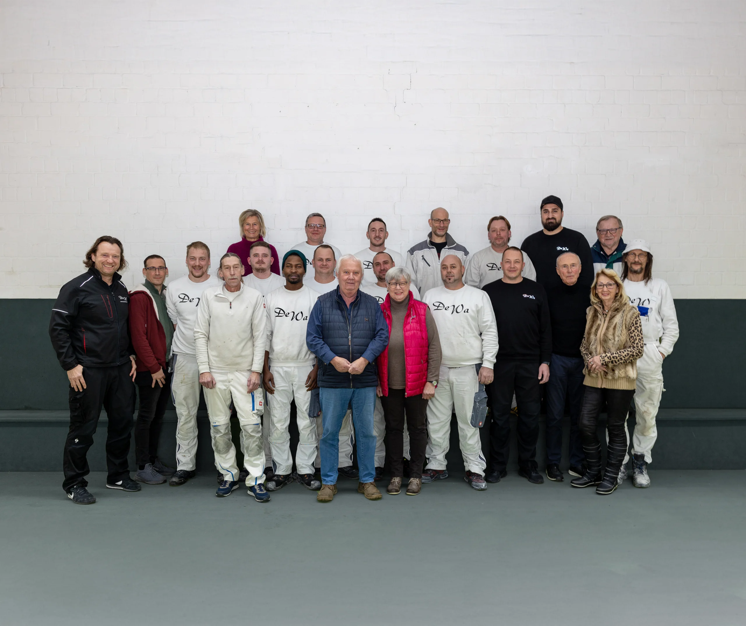 Eine Gruppe von etwa 20 Personen posiert in Arbeits- und Freizeitkleidung vor einer weißen Ziegelwand.